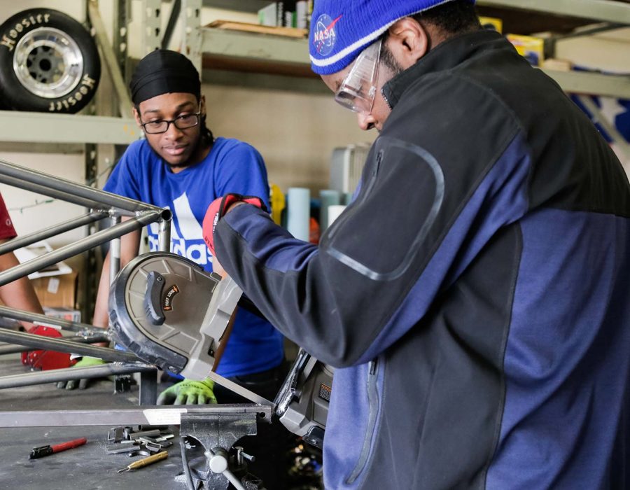 Mechanical engineering sophomore Adolf Frederic watches on as Burton makes small cuts for a suspension piece, March 21.